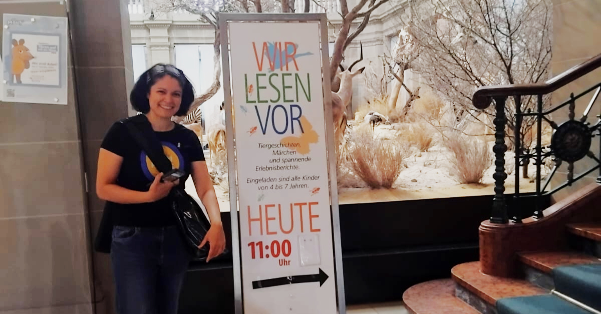 Kathrin Rosi Würtz steht in der Eingangshalle im Museum Koenig Bonn vor dem Plakat zur Veranstaltung "Wir lesen vor".